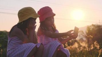 twee zussen zitten op de rails en maken samen met hun smartphones selfies bij zonsondergang. Aziatische zussen genieten ervan om samen tijd door te brengen op vakantie. video