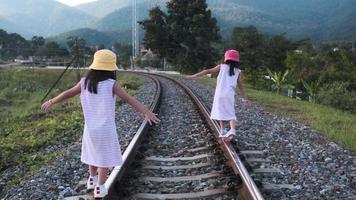 twee schattige aziatische meisjes die 's avonds op het spoor balanceren met hun armen uitgestrekt op het platteland tegen de bergen.