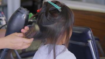 une petite fille asiatique se fait sécher les cheveux dans un salon de beauté par un coiffeur. le coiffeur fait des coiffures pour de jolies petites filles. video