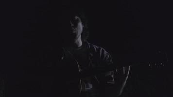 stilig lockig asiatisk man spelar gitarr och sjunger vid sjön på natten på semestern. video