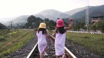 Two cute Asian girls running together on railroad tracks in the countryside against the mountains in the evening. video