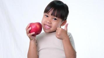 niña feliz con manzana. Linda niña asiática comiendo manzana orgánica sobre fondo blanco en el estudio. nutrición saludable para niños pequeños. video