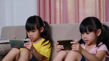 Two sisters are playing online games on their smartphones sitting on the sofa at home. Modern communication and gadget addiction concept. Two kids with gadgets.