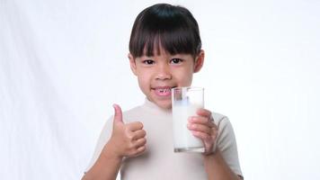 Cute Asian little girl holding a glass of milk and showing thumbs up on white background in studio. Healthy nutrition for small children.