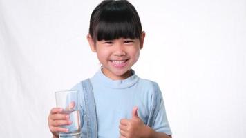 schattig klein Aziatisch meisje met een glas water en duimen opdagen op een witte achtergrond in de studio. goede gezonde gewoonte voor kinderen. gezondheidszorg concept video