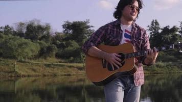 Schöner, lockiger asiatischer Mann, der im Urlaub Gitarre spielt und am See bei Sonnenuntergang singt. video