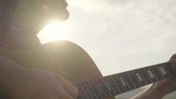 Schöner, lockiger asiatischer Mann, der im Urlaub Gitarre spielt und am See bei Sonnenuntergang singt.