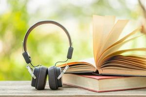Book and black headphone on wooden table with abstract green nature blur background. Reading and education concept