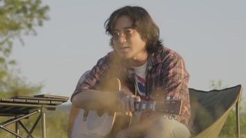 Handsome curly haired Asian man playing guitar and singing by the lake at sunset on vacation. video