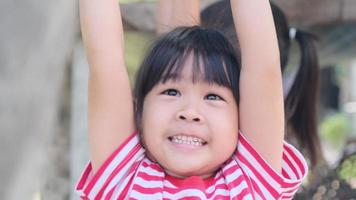menina asiática bonitinha pendura uma barra de madeira com as mãos para se exercitar no playground do quintal. criança ativa pendura na barra com as duas mãos.