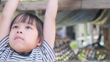söt asiatisk liten flicka hänger en trästång med händerna för träning på bakgårdens lekplats. aktivt barn hänger på baren med två händer. video