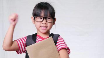 Smiling schoolgirl wearing summer outfit with backpack holding books on white background in studio. Back to school concept video