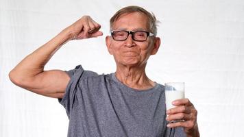 gezonde oude man die een glas melk vasthoudt terwijl hij zijn spieren laat zien en trots lacht op een witte achtergrond in de studio. video