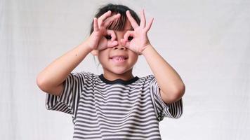 Cute little girl making okay gesture with hands on eyes and looking through fingers over a white studio background. video
