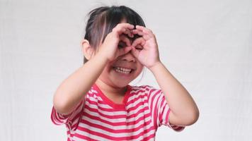 Cute little girl making funny faces wearing fingering glasses, shaped like looking through binoculars over a white studio background. video