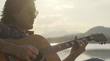 knappe aziatische man met krullend haar die gitaar speelt en zingt bij het meer bij zonsondergang op vakantie. video