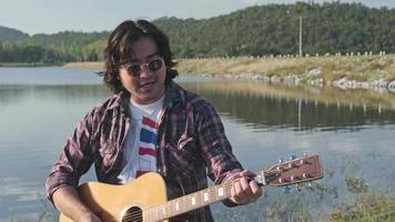 bel homme asiatique aux cheveux bouclés jouant de la guitare et chantant au bord du lac au coucher du soleil en vacances. video