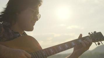 Handsome curly haired Asian man playing guitar and singing by the lake at sunset on vacation. video