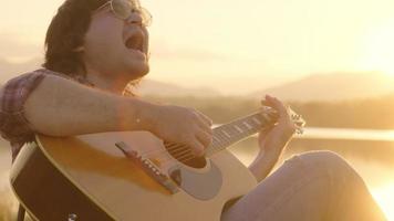 Schöner, lockiger asiatischer Mann, der im Urlaub Gitarre spielt und am See bei Sonnenuntergang singt. video