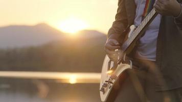 Handsome curly haired Asian man playing guitar and singing by the lake at sunset on vacation.