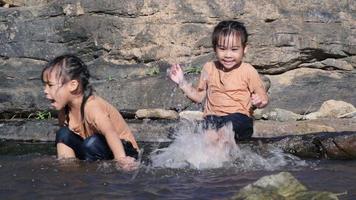 petite fille asiatique jouant dans le ruisseau de la forêt avec sa sœur. loisirs actifs avec les enfants sur la rivière en été. video