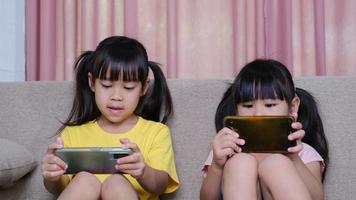 dos hermanas están jugando juegos en línea en sus teléfonos inteligentes sentadas en el sofá de casa. concepto moderno de comunicación y adicción a los gadgets. dos niños con aparatos.