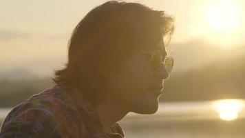 Handsome curly haired Asian man playing guitar and singing by the lake at sunset on vacation. video