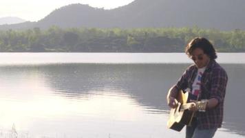 knappe aziatische man met krullend haar die gitaar speelt en zingt bij het meer bij zonsondergang op vakantie. video