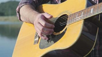 knappe aziatische man met krullend haar die gitaar speelt en zingt bij het meer bij zonsondergang op vakantie. video