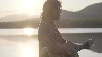 homem asiático de cabelos cacheados bonito tocando violão e cantando à beira do lago ao pôr do sol de férias.