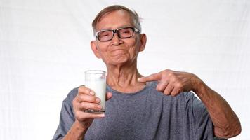 gesunder alter Mann, der ein Glas Milch hält und auf ein Glas auf weißem Hintergrund im Studio zeigt.