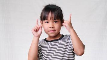 Cute little girl pointing her finger up to empty place and looking at the camera over a white studio background. video