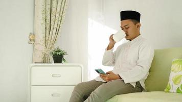 Asian Muslim man finishes worship enjoying a drink while playing on his cellphone photo