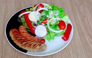 salchichas fritas con ensalada, verduras, tomate cherry, cebolla, pimentón dulce, pepino, en un plato blanco y negro. mesa de madera. Receta de dieta cetogénica o atkins. bajo en carbohidratos, alto en grasas. foto