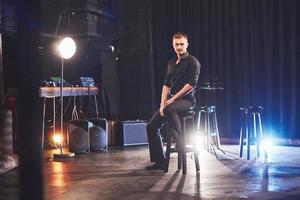 Magic look. Handsome young man in black clothes looking at camera while sitting on chair near the in dark room with light photo