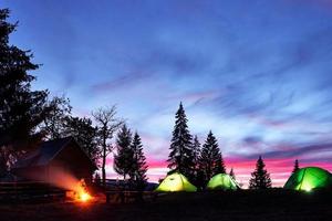 campamento nocturno. el turista descansa en una fogata cerca de una carpa iluminada y una casa de madera bajo un increíble cielo nocturno lleno de estrellas y vía láctea foto