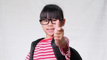 Happy schoolgirl wearing casual outfit with backpack showing thumbs up gesture on white background in studio. Back to school concept video