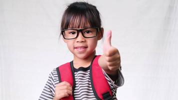 Happy schoolgirl wearing casual outfit with backpack showing thumbs up gesture on white background in studio. Back to school concept video