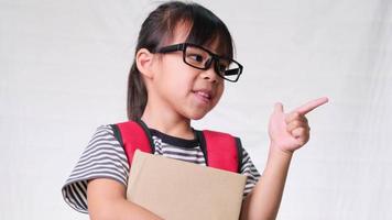 Smiling schoolgirl wearing casual outfit with backpack holding books and pointing empty space sideways on white background in studio. Back to school concept video
