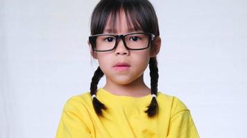 garota irritada com óculos e uma camiseta amarela fica com os braços cruzados contra um fundo branco no estúdio. menina bonitinha está chateada sozinha em casa. video