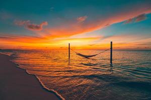 Tropical sunset beach as summer landscape panorama with beach swing or hammock. Colorful sky, sand palm tree leaves and calm sea beach banner. Couple romantic beach scene photo