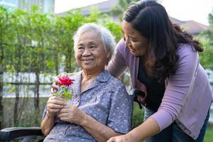 Caregiver daughter hug and help  Asian senior or elderly old lady woman holding red rose on wheelchair in park. photo