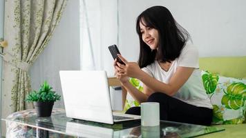mujer asiática que usa teléfono móvil mientras trabaja en casa con una computadora portátil en la silla foto
