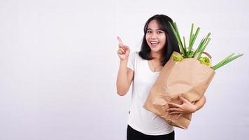 hermosa mujer asiática sosteniendo una bolsa de papel de verduras frescas muy feliz señalando con la mano y el dedo en un fondo blanco aislado foto