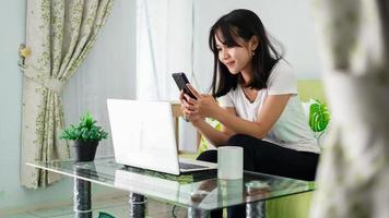 asian woman using mobile phone while working at home with laptop on chair photo