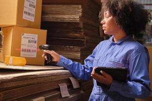 African American female worker in safety uniform using bar code scanner to check shipment orders at parcels warehouse, paper manufacture factory for the packing industry, logistic transport service. photo