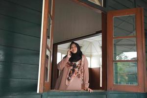 Muslim woman doing phone call while smiling and looking through window photo