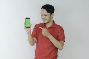 un joven asiático inteligente está feliz y sonriendo cuando muestra la pantalla verde del teléfono inteligente en el fondo del estudio foto