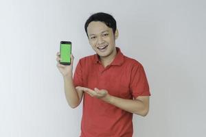 un joven asiático inteligente está feliz y sonriendo cuando muestra la pantalla verde del teléfono inteligente en el fondo del estudio foto