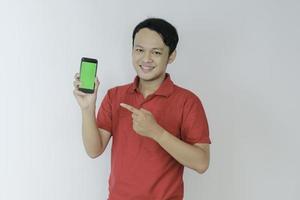 un joven asiático inteligente está feliz y sonriendo cuando muestra la pantalla verde del teléfono inteligente en el fondo del estudio foto